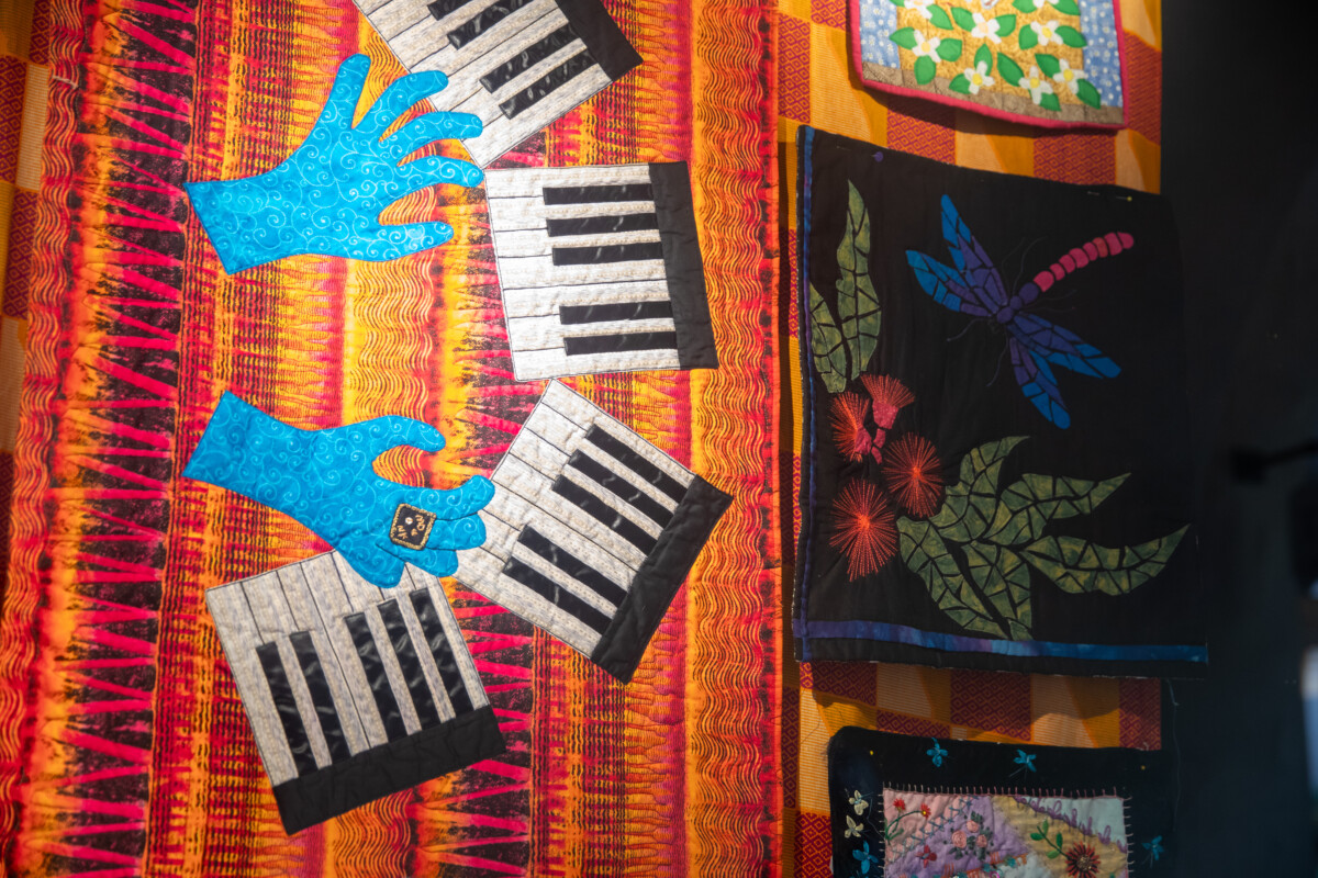 Art quilt on display depicting hands playing piano and a dragonfly with fruit