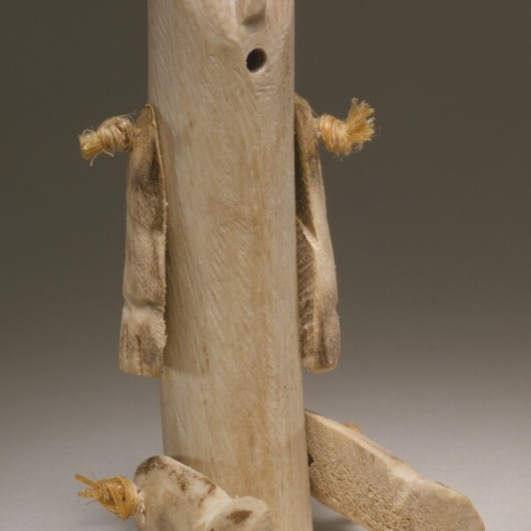 This Inuit Doll is made from a caribou antler. The body of the doll is cylindrical. Two arms and two legs are attached to body. The arms are positioned straight down to the ground. The legs are positioned out in front as if the doll was seated. The top of the caribou antler has been rounded to create a face. The mouth and eyes are open holes, while the nose was carved..