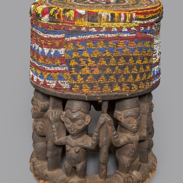 This stool was carved from a single block of wood. Its openwork base features caryatid figures (female figures, used as a pillar to support the entablature of a Greek or Greek-style building). The top of the stool is decorated with beads which indicates it was intended more for display at public events and ceremonial occasions rather than for everyday seating.