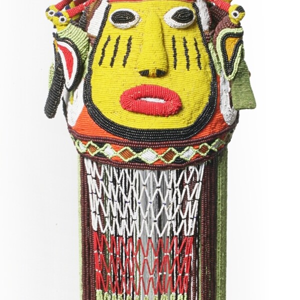 This crown is beaded from top to bottom. On top of the crown sits a bead bird and below the bird is a human face. Long strands of beads are below the face.