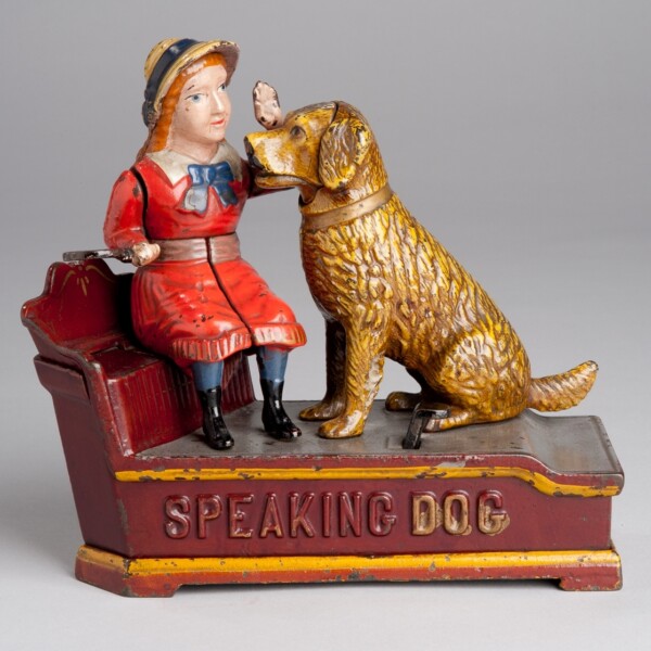 This cast iron mechanical toy bank features a young girl sitting on the bench petting her dog. The dog is in a seated position and is the same height as the girl sitting on the bench. The girl is wearing a read dress with a blue bow and possibly a straw hat. Underneath the girl and the dog are the words "speaking dog".