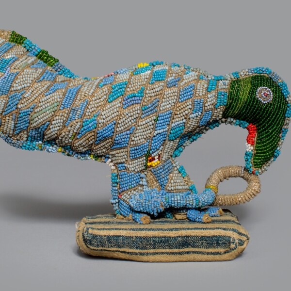 Only seven inches tall, this figure comprised of glass beads, cotton, and wood depicts a bird attacking a snake with its beak. The body of the bird is beaded with blue, green, and cream beads in a checkered diamond pattern. The head of the bird uses green beads. The snake is white beads.
