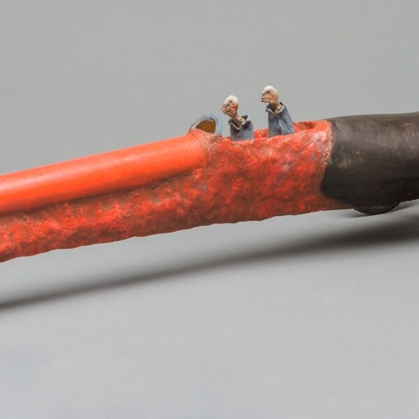 This piece is a a long skinny vehicle with two small wheels at the front and two larger wheels on the back. The front three quarters of the vehicle is red/orange and the rest is black. Two figures operating the vehicle from inside the middle area.