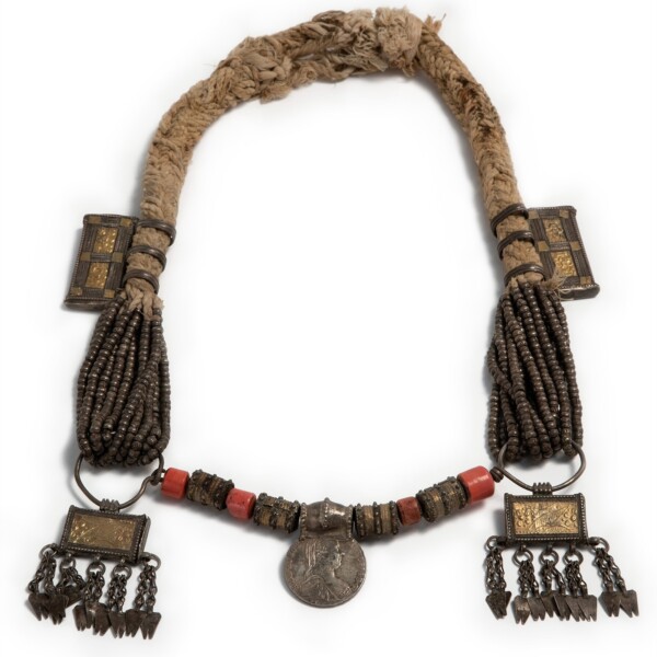 This necklace is a mix of materials such as cotton rope, glass and silver. In the center is a silver medallion and to either side of the medallion are glass beads. Some of the glass beads are made to look like coral..