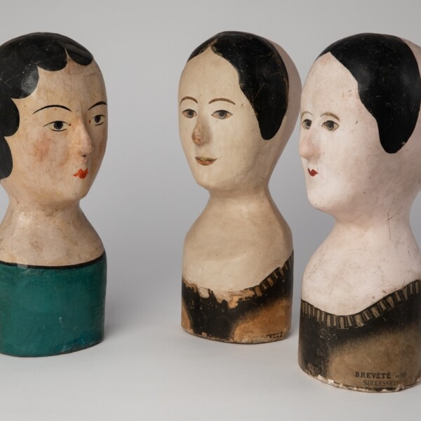 There are three milliner's store head forms. The two on the right face the one on the left.  The two on the right have short black hair. The one on the left has long black hair. They are all light complexioned.
