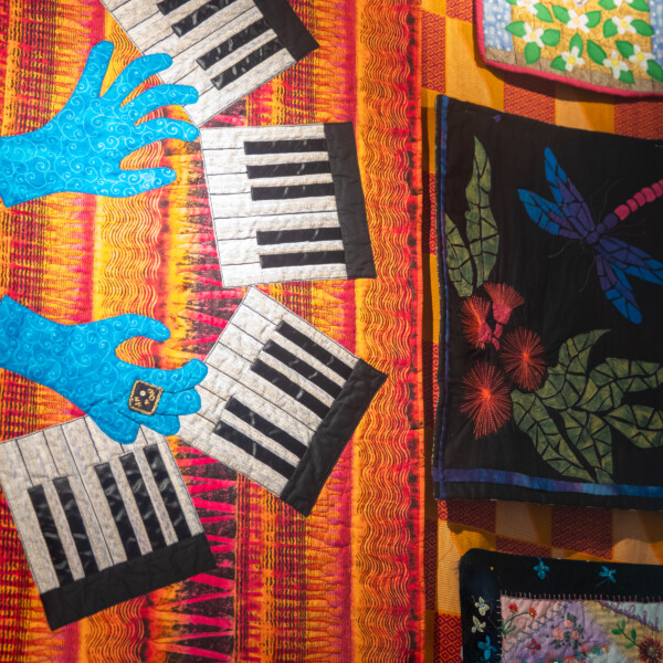 Art quilt on display depicting hands playing piano and a dragonfly with fruit